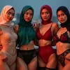 Four women in hijabs and lingerie pose on a rooftop with city lights in the background.